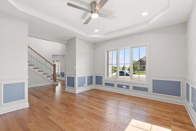 empty room featuring a raised ceiling, stairway, light wood-style flooring, ceiling fan, and recessed lighting