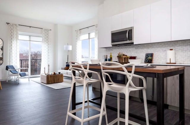 kitchen featuring dark wood-type flooring, white cabinets, backsplash, stainless steel microwave, and a kitchen bar