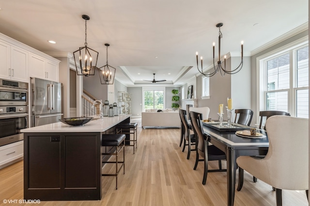 kitchen with appliances with stainless steel finishes, light wood-type flooring, a kitchen island with sink, ceiling fan with notable chandelier, and a tray ceiling