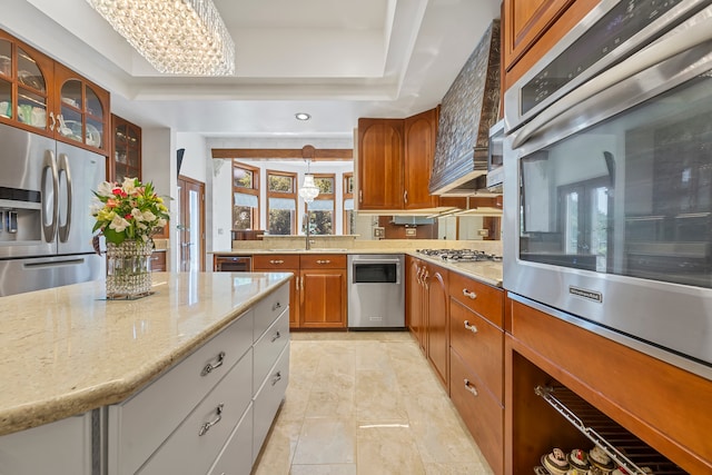 kitchen with appliances with stainless steel finishes, light stone counters, custom range hood, a tray ceiling, and sink