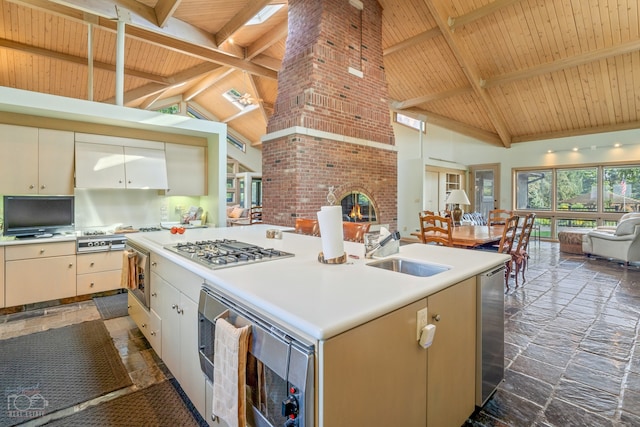 kitchen featuring a brick fireplace, a kitchen island with sink, wood ceiling, appliances with stainless steel finishes, and beamed ceiling