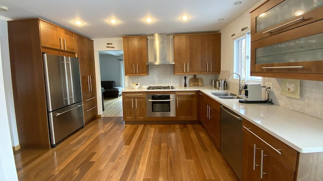 kitchen with brown cabinetry, wood finished floors, a sink, stainless steel appliances, and wall chimney exhaust hood