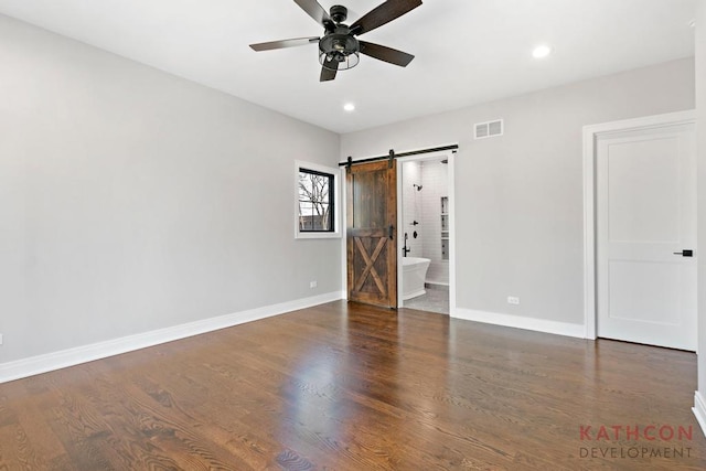 unfurnished bedroom featuring a barn door, dark hardwood / wood-style flooring, ceiling fan, and ensuite bathroom