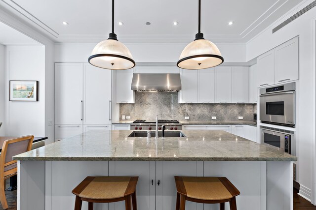 kitchen featuring tasteful backsplash, crown molding, wall chimney range hood, double oven, and a sink