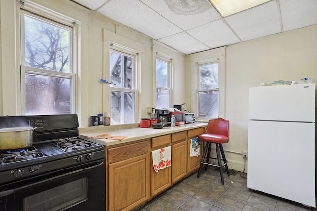 kitchen with a paneled ceiling, white refrigerator, black gas range, and dark tile patterned floors