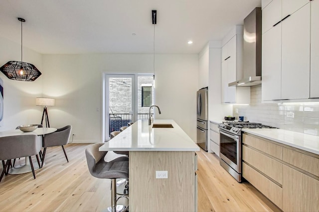 kitchen with sink, hanging light fixtures, appliances with stainless steel finishes, light hardwood / wood-style flooring, and wall chimney exhaust hood