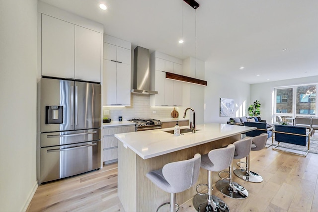 kitchen with hanging light fixtures, light wood-type flooring, wall chimney range hood, stainless steel appliances, and a kitchen island with sink