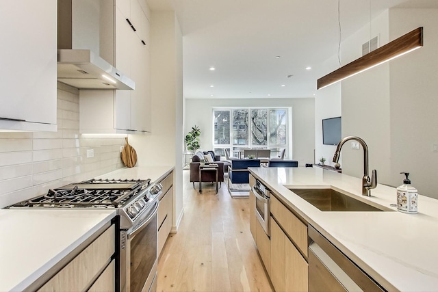 kitchen with light brown cabinetry, light wood-type flooring, wall chimney range hood, stainless steel appliances, and sink