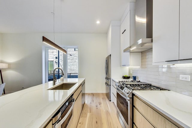 kitchen with sink, appliances with stainless steel finishes, light stone countertops, wall chimney exhaust hood, and light wood-type flooring