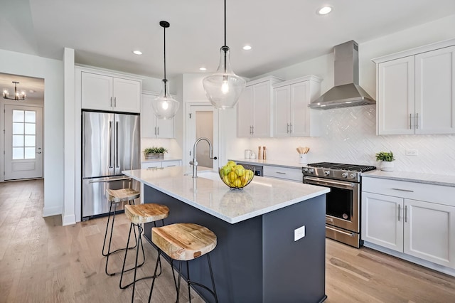 kitchen with appliances with stainless steel finishes, wall chimney exhaust hood, a kitchen island with sink, and white cabinets