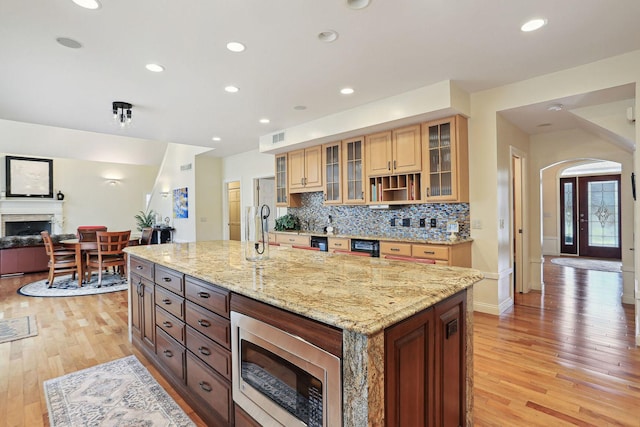 kitchen featuring light stone counters, light wood-type flooring, a large island with sink, stainless steel microwave, and decorative backsplash