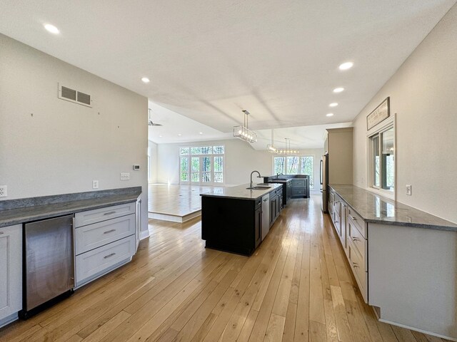 kitchen featuring an inviting chandelier, an island with sink, light wood-type flooring, pendant lighting, and dark stone countertops