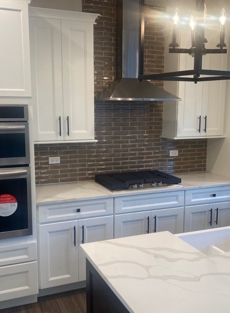 kitchen with light stone countertops, backsplash, stainless steel appliances, white cabinets, and wall chimney range hood