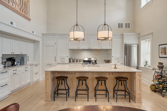 kitchen featuring pendant lighting, a towering ceiling, backsplash, a center island with sink, and stainless steel refrigerator