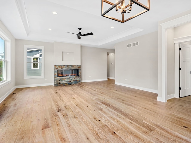 unfurnished living room featuring a healthy amount of sunlight, light hardwood / wood-style floors, a tray ceiling, a fireplace, and ceiling fan with notable chandelier