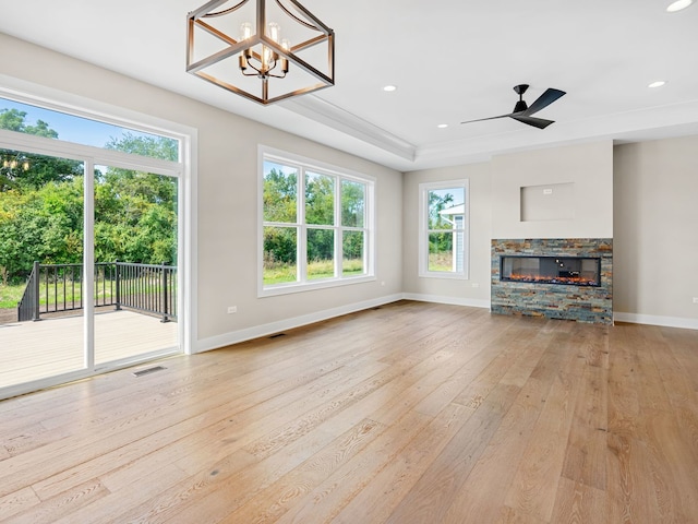 unfurnished living room featuring ceiling fan with notable chandelier, light hardwood / wood-style floors, a stone fireplace, and a tray ceiling