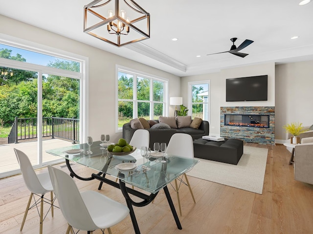 dining space featuring a fireplace, light wood-type flooring, a tray ceiling, and ceiling fan with notable chandelier