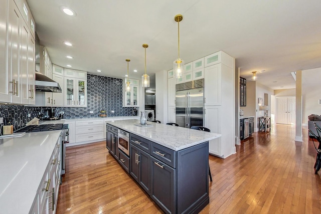 kitchen featuring built in appliances, white cabinetry, a center island with sink, and hanging light fixtures