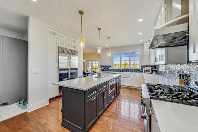 kitchen featuring a kitchen island with sink, exhaust hood, built in appliances, decorative light fixtures, and white cabinets