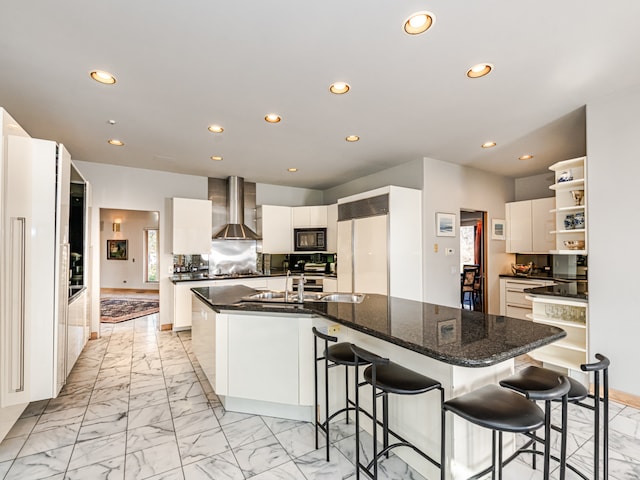 kitchen with built in appliances, wall chimney exhaust hood, an island with sink, and white cabinets