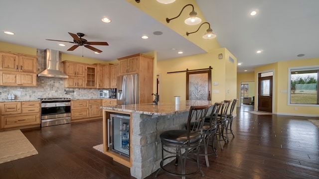 kitchen featuring a barn door, appliances with stainless steel finishes, a breakfast bar area, wall chimney range hood, and dark hardwood / wood-style flooring