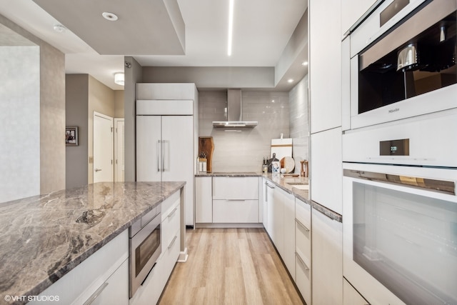 kitchen featuring wall chimney range hood, light wood-type flooring, white cabinetry, built in appliances, and light stone counters