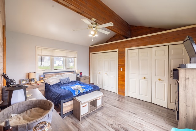 bedroom with two closets, light wood-type flooring, wooden walls, ceiling fan, and lofted ceiling with beams