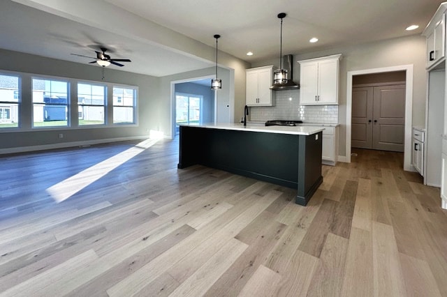 kitchen with wall chimney range hood, ceiling fan, white cabinetry, a kitchen island with sink, and light wood-type flooring