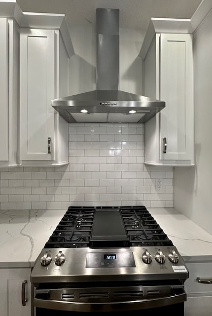 kitchen featuring wall chimney range hood, gas range, white cabinets, and light stone countertops