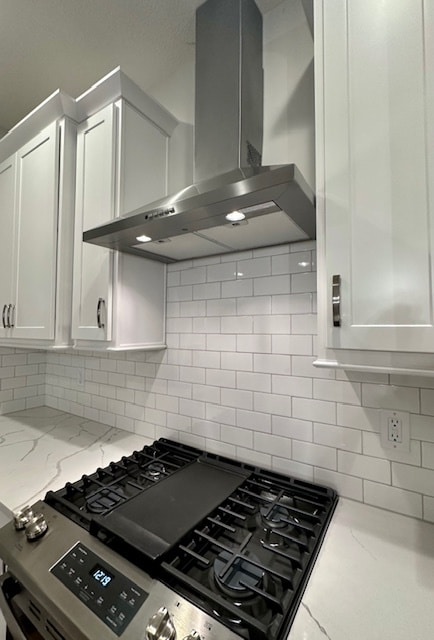 kitchen featuring wall chimney range hood, white cabinetry, backsplash, gas stove, and light stone counters