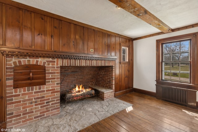 unfurnished living room featuring a textured ceiling, beamed ceiling, wood-type flooring, a brick fireplace, and radiator heating unit