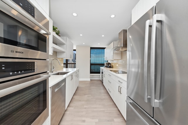 kitchen with appliances with stainless steel finishes, wall chimney exhaust hood, sink, white cabinets, and light hardwood / wood-style floors