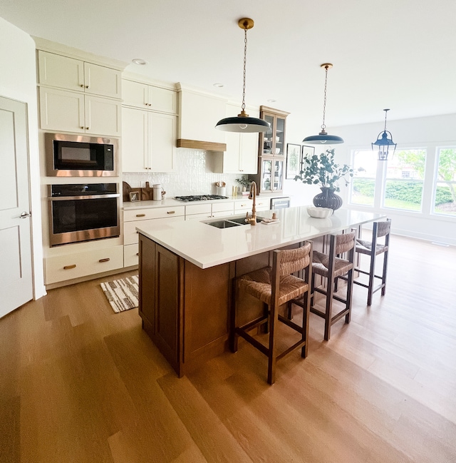 kitchen featuring appliances with stainless steel finishes, a center island with sink, light hardwood / wood-style flooring, tasteful backsplash, and hanging light fixtures