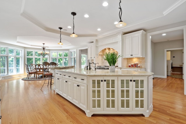 kitchen featuring light wood-type flooring, white cabinets, an island with sink, a tray ceiling, and pendant lighting