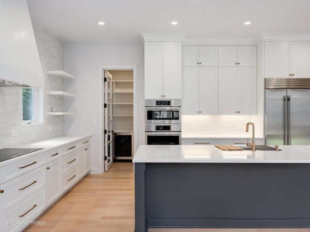 kitchen with backsplash, stainless steel appliances, custom range hood, a center island with sink, and light hardwood / wood-style floors