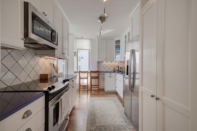 kitchen with stainless steel appliances, dark countertops, glass insert cabinets, white cabinetry, and light wood-type flooring