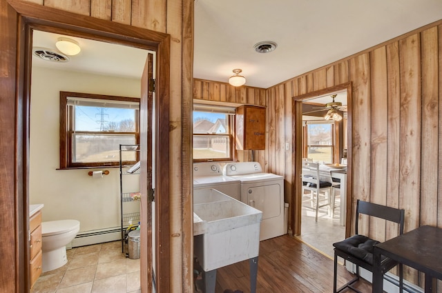 clothes washing area with wood walls, a baseboard radiator, light hardwood / wood-style floors, ceiling fan, and washer and dryer