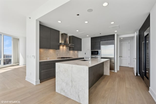 kitchen featuring a center island with sink, wall chimney exhaust hood, built in appliances, backsplash, and sink