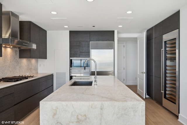 kitchen featuring wall chimney range hood, light hardwood / wood-style flooring, stainless steel appliances, backsplash, and a center island with sink