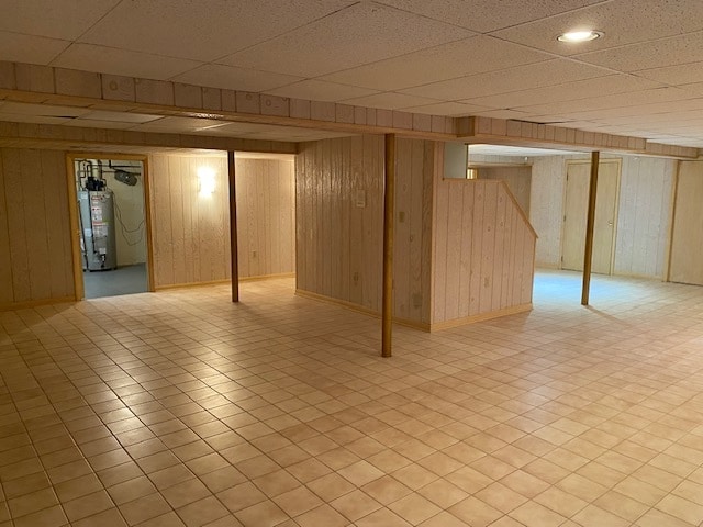 basement with wood walls, a paneled ceiling, water heater, and light tile patterned floors