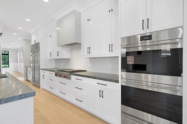 kitchen with wall chimney exhaust hood, white cabinetry, light hardwood / wood-style flooring, stainless steel appliances, and backsplash