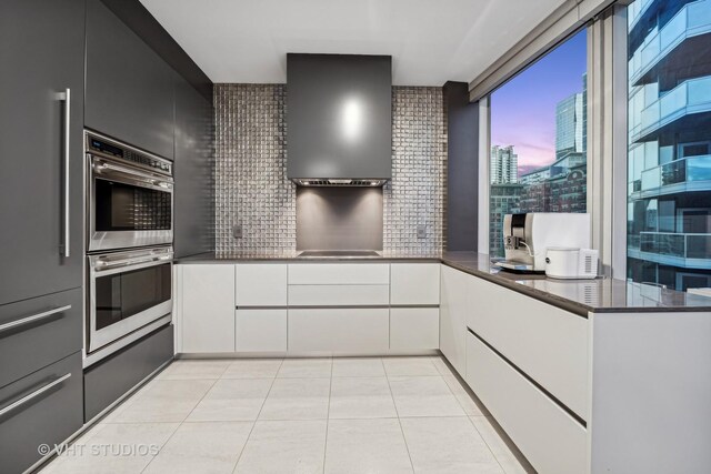 kitchen with light tile patterned floors, decorative backsplash, white cabinetry, stainless steel double oven, and wall chimney exhaust hood