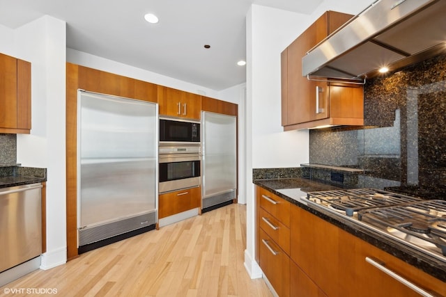 kitchen featuring dark stone counters, built in appliances, decorative backsplash, range hood, and light hardwood / wood-style floors