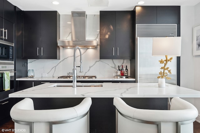 kitchen with a kitchen island with sink, backsplash, wall chimney range hood, a breakfast bar area, and appliances with stainless steel finishes