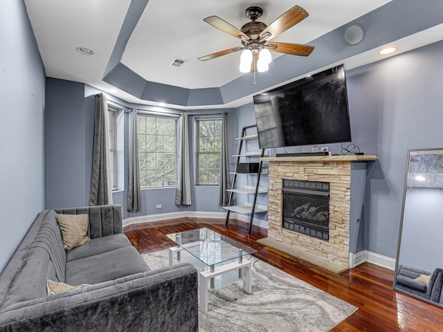 living room with dark hardwood / wood-style flooring, ceiling fan, a stone fireplace, and a tray ceiling