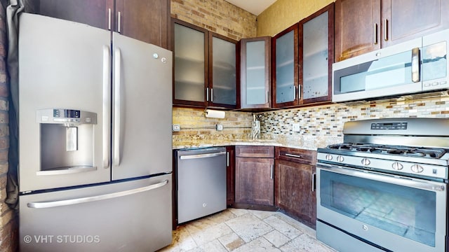 kitchen with light stone counters, a sink, appliances with stainless steel finishes, decorative backsplash, and glass insert cabinets