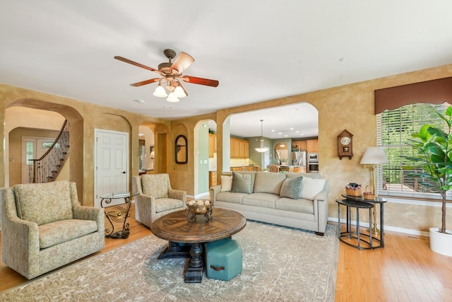 living area with arched walkways, light wood-type flooring, ceiling fan, baseboards, and stairway