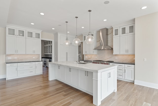 kitchen with white cabinets, wall chimney exhaust hood, pendant lighting, light wood-type flooring, and a kitchen island with sink