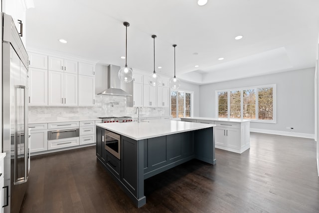 kitchen featuring decorative light fixtures, a kitchen island with sink, wall chimney range hood, built in appliances, and dark hardwood / wood-style floors