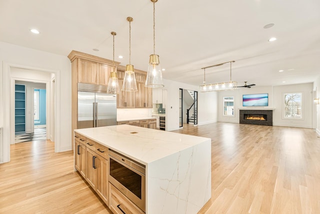 kitchen with light brown cabinetry, light wood-type flooring, built in appliances, a warm lit fireplace, and a kitchen island